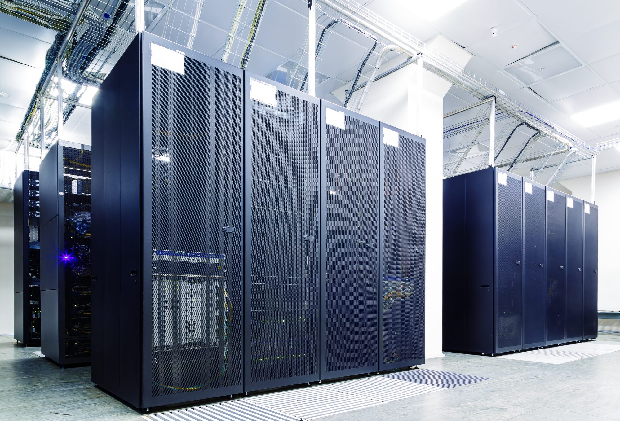 Data Centers Rows of black server racks in a well-lit data center with visible cables and cooling vents on the floor.